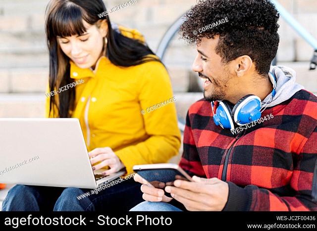 Man with digital tablet smiling while sitting by friend using laptop outdoors