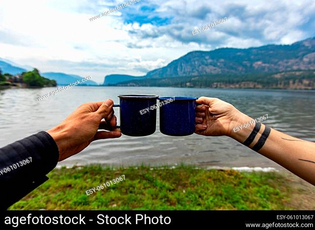 Selective focus of two different people clinching their cups of coffee, or tea to each other with a nature background with a lake and mountains