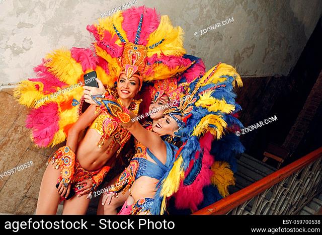 Woman in brazilian samba carnival costume with colorful feathers plumage with mobile phone take selfie in old entrance with big window