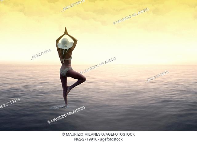 Woman with a white hat standing on the water in the sun salutation yoga pose, on background sky with colored clouds by a warm sunset
