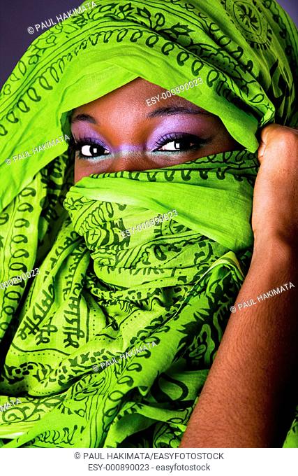 The face of an innocent beautiful young African-American woman covering her mouth showing only her eyes with green headwrap and purple-green makeup, isolated