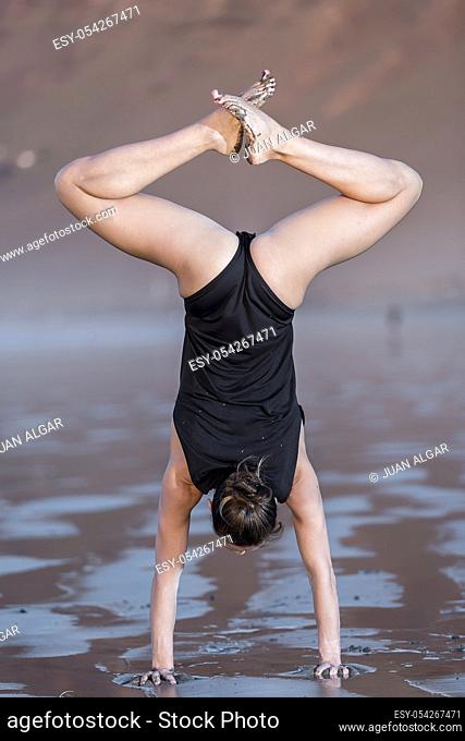 Back view of slim woman in black bodysuit doing handstand with crossed legs on wet sand of beach