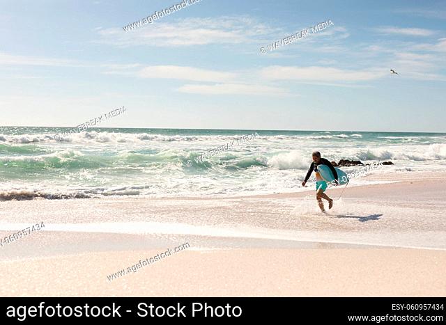 Retired biracial senior man running with surfboard on shore at beach against sky during sunny day