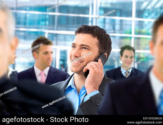 Happy businessman talking on mobile standing in crowd in office lobby