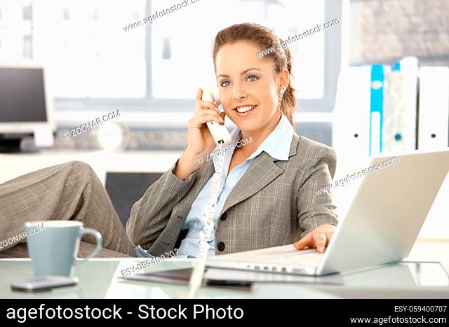Attractive businesswoman sitting at desk, talking on phone, having laptop, smiling