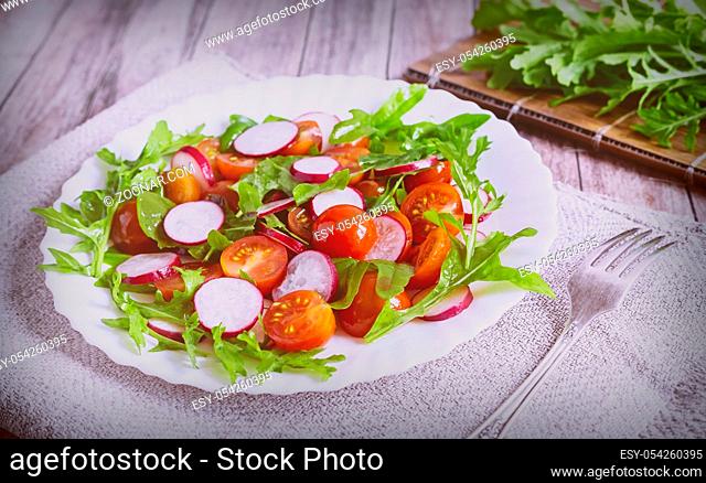 On the table on a white plate of radish salad, tomato, arugula, near a bunch of greens