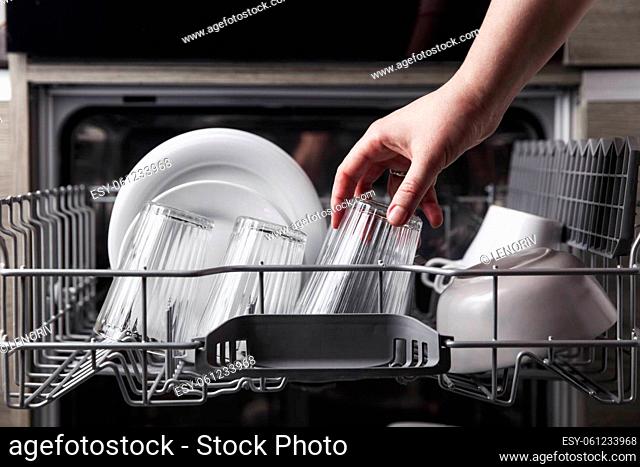 Close-up of female hand loading dished to, empty out or unloading from open automatic stainless built-in dishwasher machine with clean utensils inside in modern...