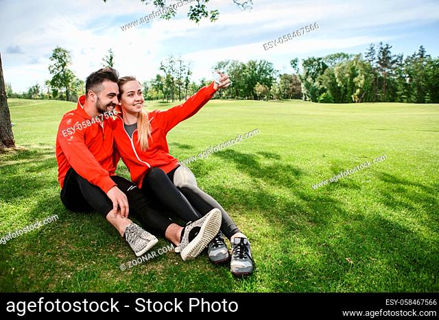 Sport man and woman making selfies in green park or forest while sitting on green grass. Happy couple hugging and posing for camera