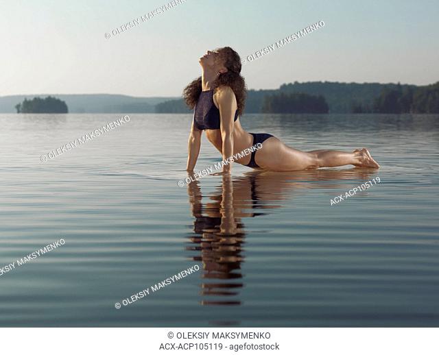 Young woman practicing Hatha yoga on a platform in water on the lake in the morning. Yoga Upward Facing Dog, Cobra variation posture, Urdhva Mukha Svanasana