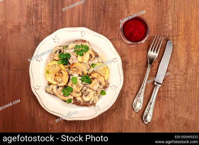 Overhead photo of scallopine di vitello, veal scallopini, with mushroom sauce, lemons, and parsley, shot from above on dark rustic texture with glass of red...