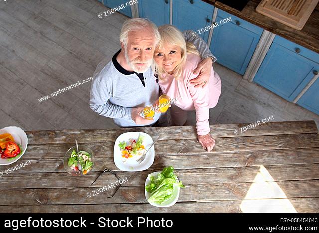 Photo of elderly couple hugging in their kitchen. Picture shot from above of two aged people in hug next to wooden table with food on it at their home