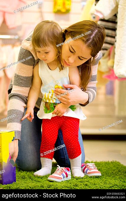 Young attractive mother with her daughter looking at the toy in children's store, telephoto shot