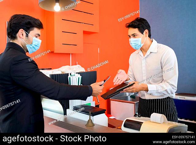 The cashier in the apron and face mask hands a receipt to a caucasian customer. A young businessman in a suit pay a service fee through Mobile applications