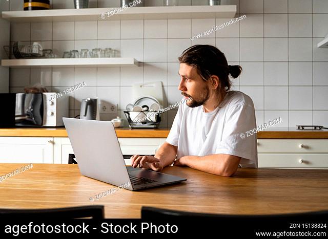 Freelancer working from home on the kitchen and using laptop. Bearded man working with a laptop and reading good news. Handsome successful self entrepreneur...