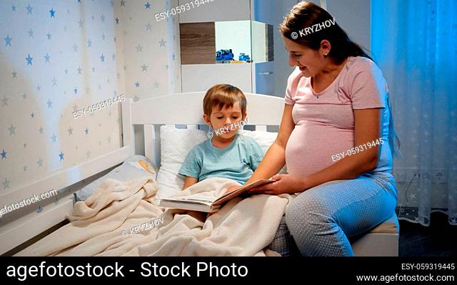 Beautiful pregnant mother readng story book to her older son lying in bed at night before going to sleep. Parents reading to children