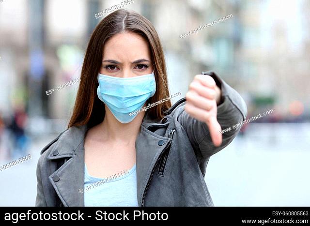 Front view portrait of serious girl doing thumbs down gesture with protective mask on city street