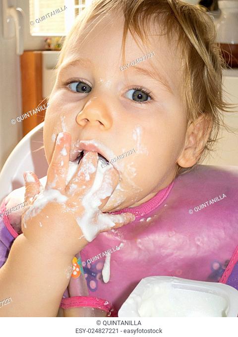 blonde caucasian baby 1 year eating natural yogurt with her hand in high-chair kitchen