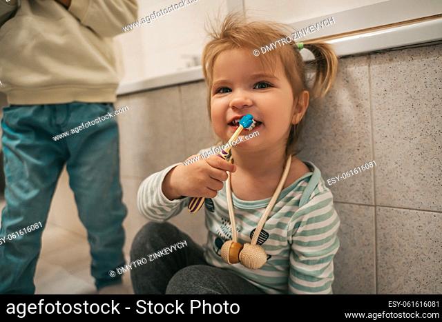 Cute female child sitting in the bathroom and brushing her teeth in the presence of her sibling