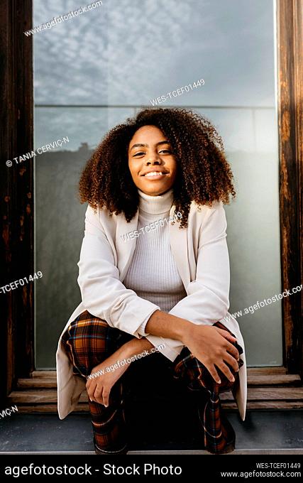 Smiling afro young woman wearing white coat sitting on window sill