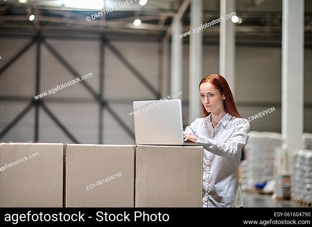 Important point. Young elegant woman standing in warehouse near stacked boxes working looking intently at laptop