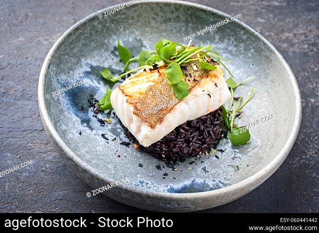 Modern style traditional fried skrei cod fish filet with portulaca lettuce and black rice served as close-up on ceramic design plate with copy space