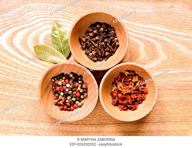 Three wooden bowls with pepper, cloves and slices of dried peppers on a light wooden table. Next to the bowl are two of dried bay leaves