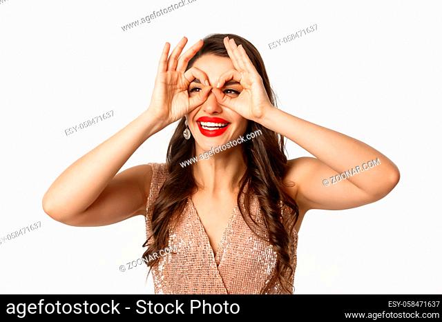 Concept of New Year celebration and winter holidays. Close-up of beautiful brunette woman in dress, red lips, making hand binoculars and staring left