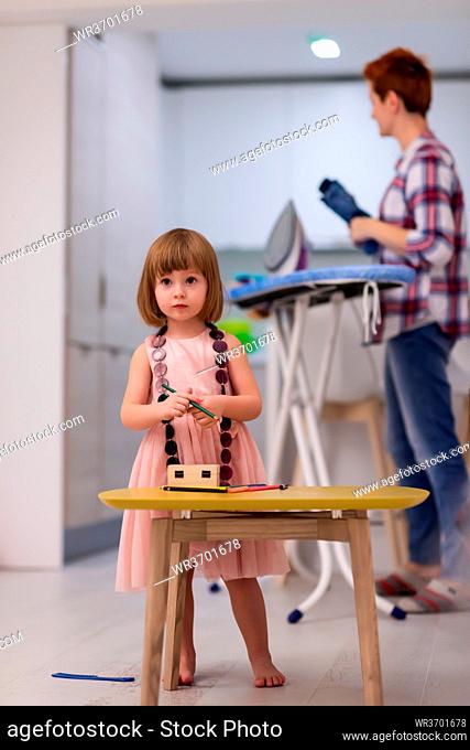 happy family spending time together at home cute little daughter in a pink dress playing and painting the jewelry box while young redhead mother ironing clothes...