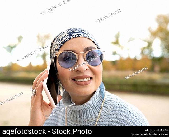 Young woman wearing headscarf and sunglasses smiling while talking on mobile phone outdoors