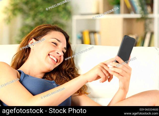 Happy woman using smart phone lying on a couch in the living room at home
