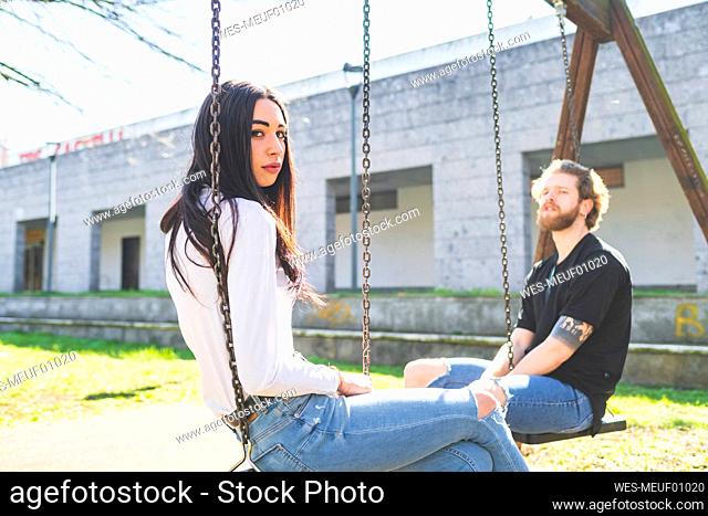 Young couple sitting on swings in park during sunny day