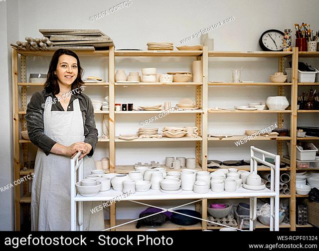 Smiling craftswoman standing with ceramics in cart at workshop