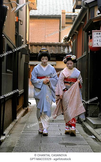 A GEIKO GEISHA, ON THE LEFT, AND A MAIKO APPRENTICE WEARING A KIMONO OBEBE HELD CLOSED BY A WIDE BELT OBI AND WOOD SANDALS OKOBO, WALKING DOWN A STREET