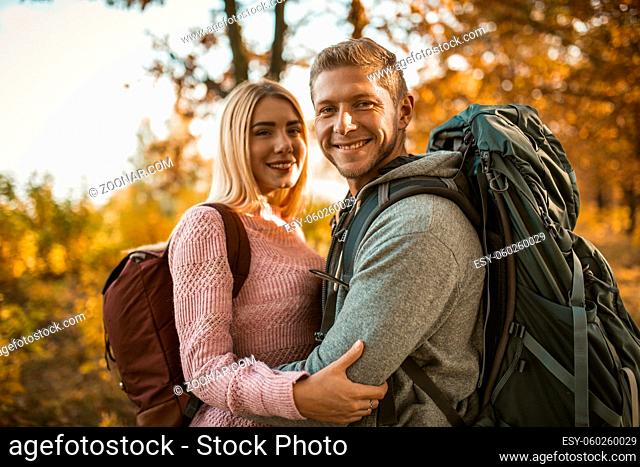 Happy couple of backpackers hugging while looking at the camera, a Caucasian couple in love toothy laughs while standing against the backdrop of colorful autumn...