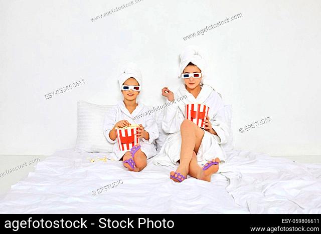 happy woman with daughter in white bathrobes and towels on heads are watching movie on tv sitting on bed