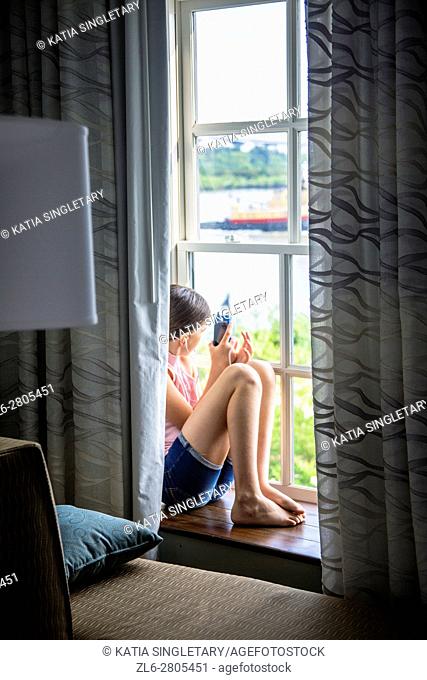 Gorgeous long hair caucasian preteen in her 10, 11, 12, 13 years old sitting on the window seal of the hotel room, . She is taking photos with her phone and...
