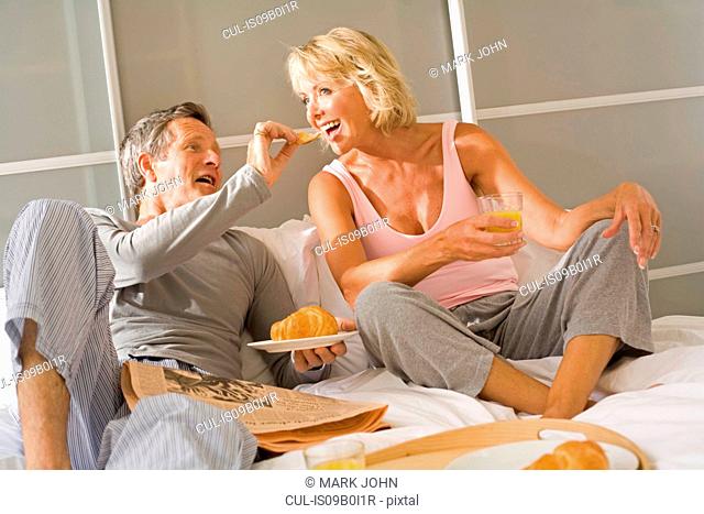 Romantic senior man feeding breakfast croissant to wife in bed