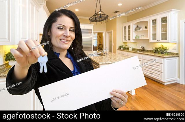 Pretty hispanic woman in kitchen holding house keys and blank white sign