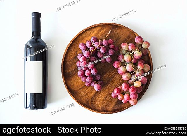 Red wine bottle, large burgundy fresh grapes on round wooden dish on white background, copy space flat lay, top view, mockup