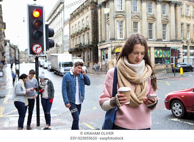 A young woman can be seen walking along a city street with a smart phone. Other young adults are in the background using smart phones