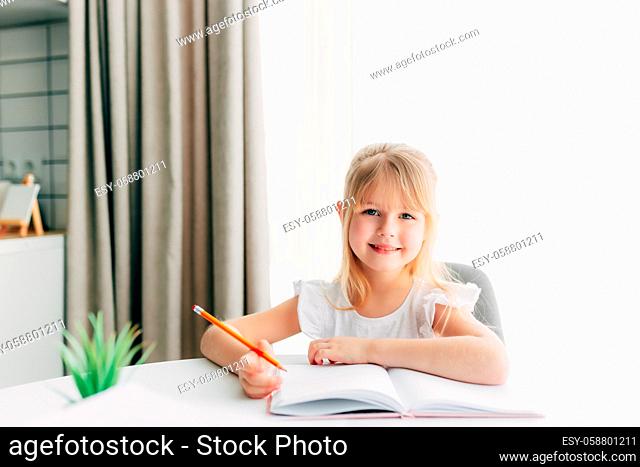 A little smiling girl sits at the table and writes in a white notebook. Education concept. Home schooling. Homework. Smiling face. High quality photo
