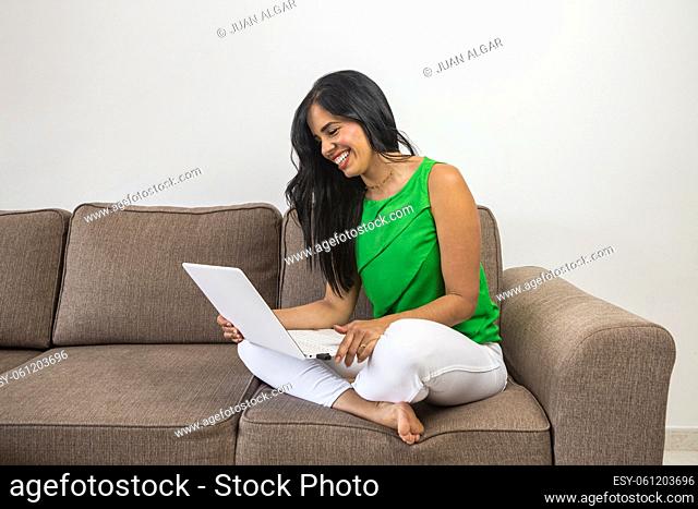 Hispanic female in casual clothes with long dark hair watching video on netbook and laughing at joke while sitting on couch in daytime at home