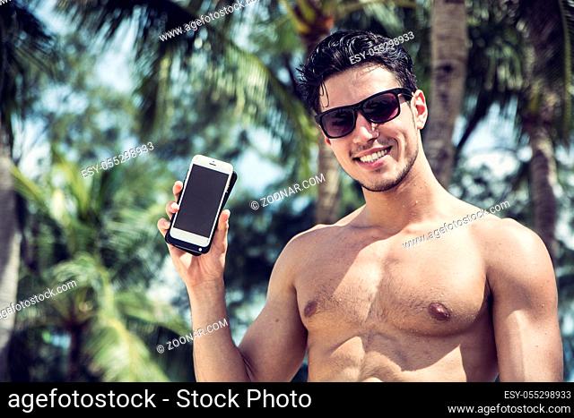 Shirtless Young Handsome Man Showing Cell Phone While Smiling to Camera, with Palm Trees Behind Him. Wearing Sunglasses