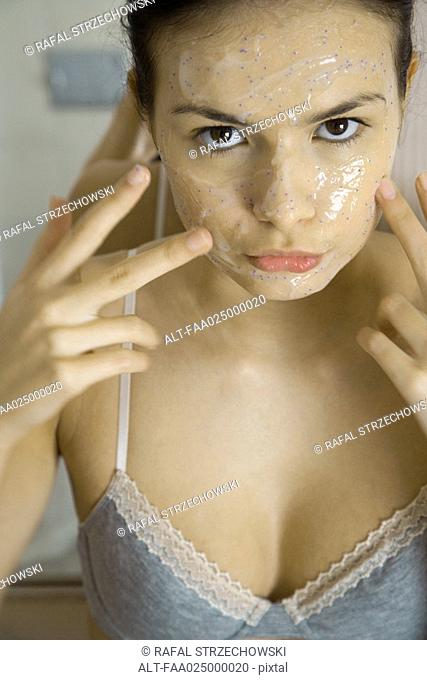 Young woman applying facial mask, looking at camera, close-up
