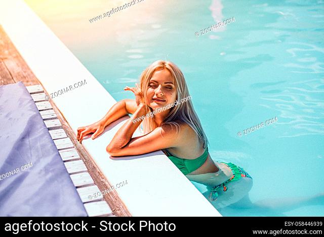 Beautiful blonde girl in hotel swimming pool in green bikini with her eyes closed with long straight hair. Girl satisfy the vacation and sitting on the corner...