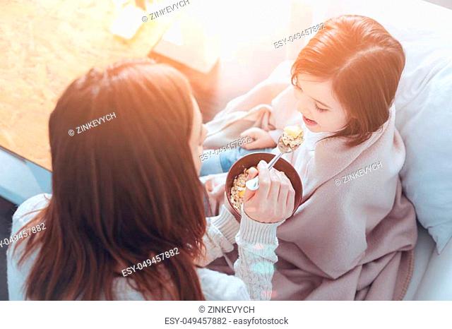 Dinner time. Kind mother holding spoon in right hand near opened mouth of her daughter and looking forward while sitting in semi position