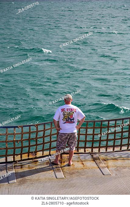 Caucasian American man with a NC State teeshirt, short and a baseball cap looking at the ocean during the crossing on the Ferry in the outer banks of North...