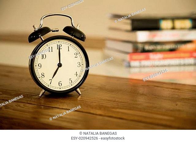 Vintage alarm clock on rustic wood table. Shows 7 o'clock. Pile of books in background