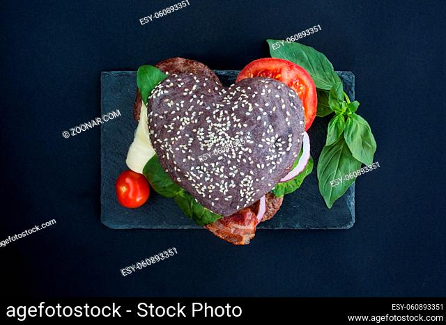 Heart shaped black hamburger on stone plate, love burger fast food concept, Valentines day surprise dinner, wooden background, top view flat lay