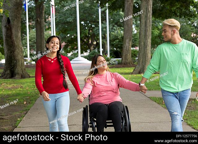 Young woman in a wheelchair holds hands with the friends that are walking down a path beside her
